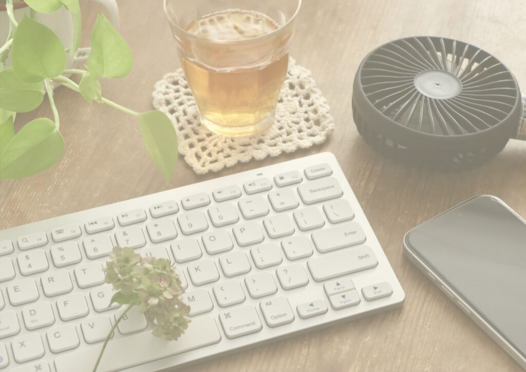 木製のデスクにキーボード、スマートフォン、アイスティー、観葉植物、携帯扇風機が置かれた在宅ワーク環境の写真。インボイス制度に関する解説記事のイメージ。